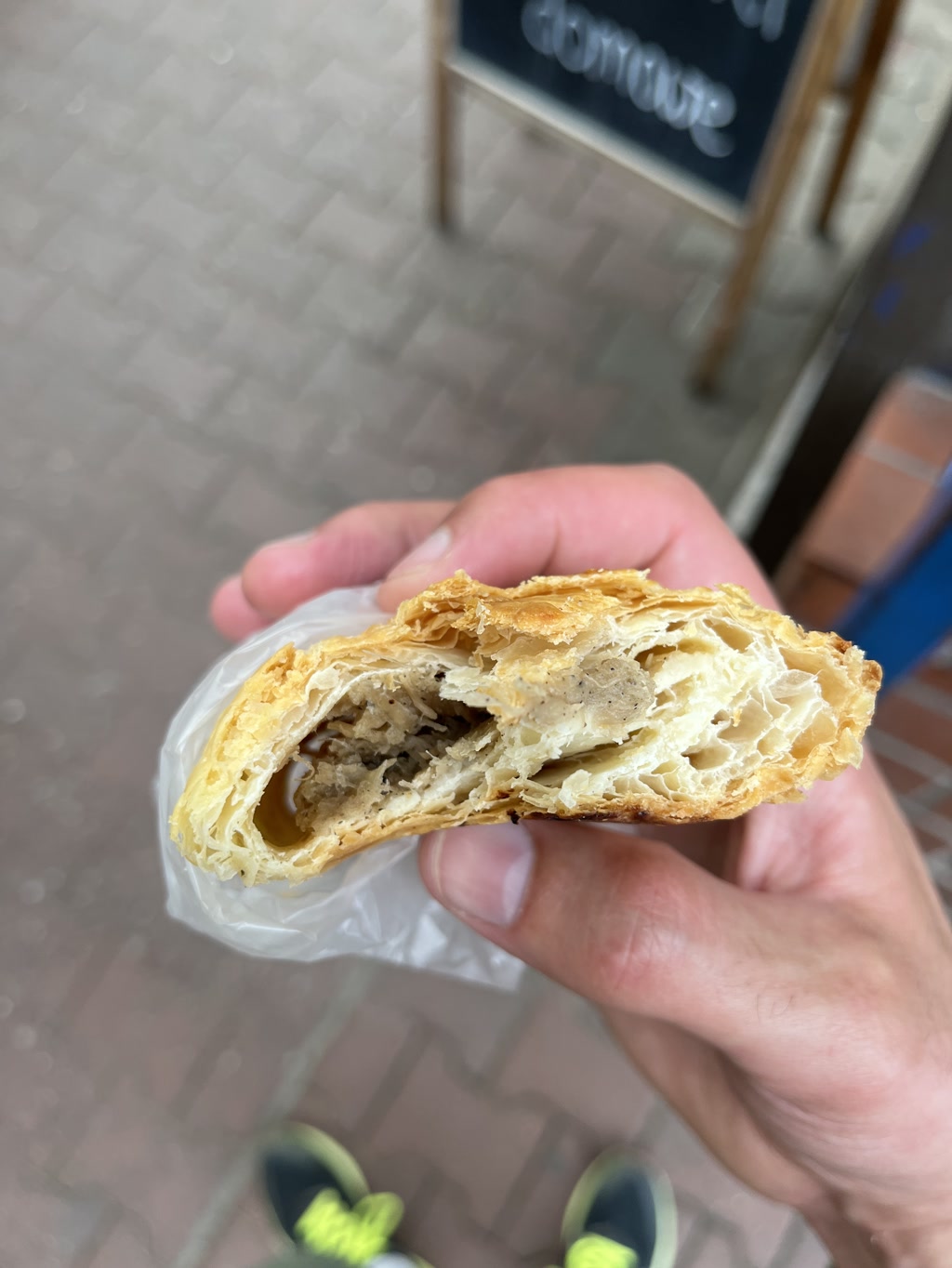 A person is holding a half-eaten savory pastry with a flaky, golden-brown crust in their hand. The pastry's layered interior is visible, filled with a ground meat mixture that appears seasoned, suggesting it might be a meat pie or a sausage roll. The hand is holding a small clear plastic bag wrapped around the bottom half of the pastry, possibly for cleanliness while eating. In the background, there is a blurred facade of a building with glass doors and a sign above them, though the text is indiscernible and not the focus of the scene.